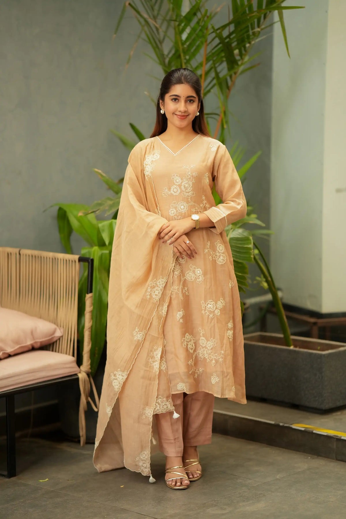 A woman models a traditional Indian outfit consisting of a beige kurta with white floral embroidery, matching trousers, and a draped dupatta, set against a modern indoor backdrop with plants and a bench.