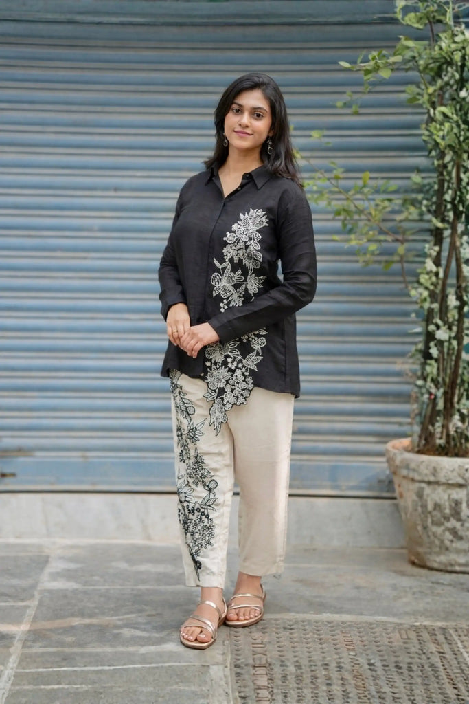 Woman wearing a black embroidered shirt and beige pants standing in front of a blue shutter.
