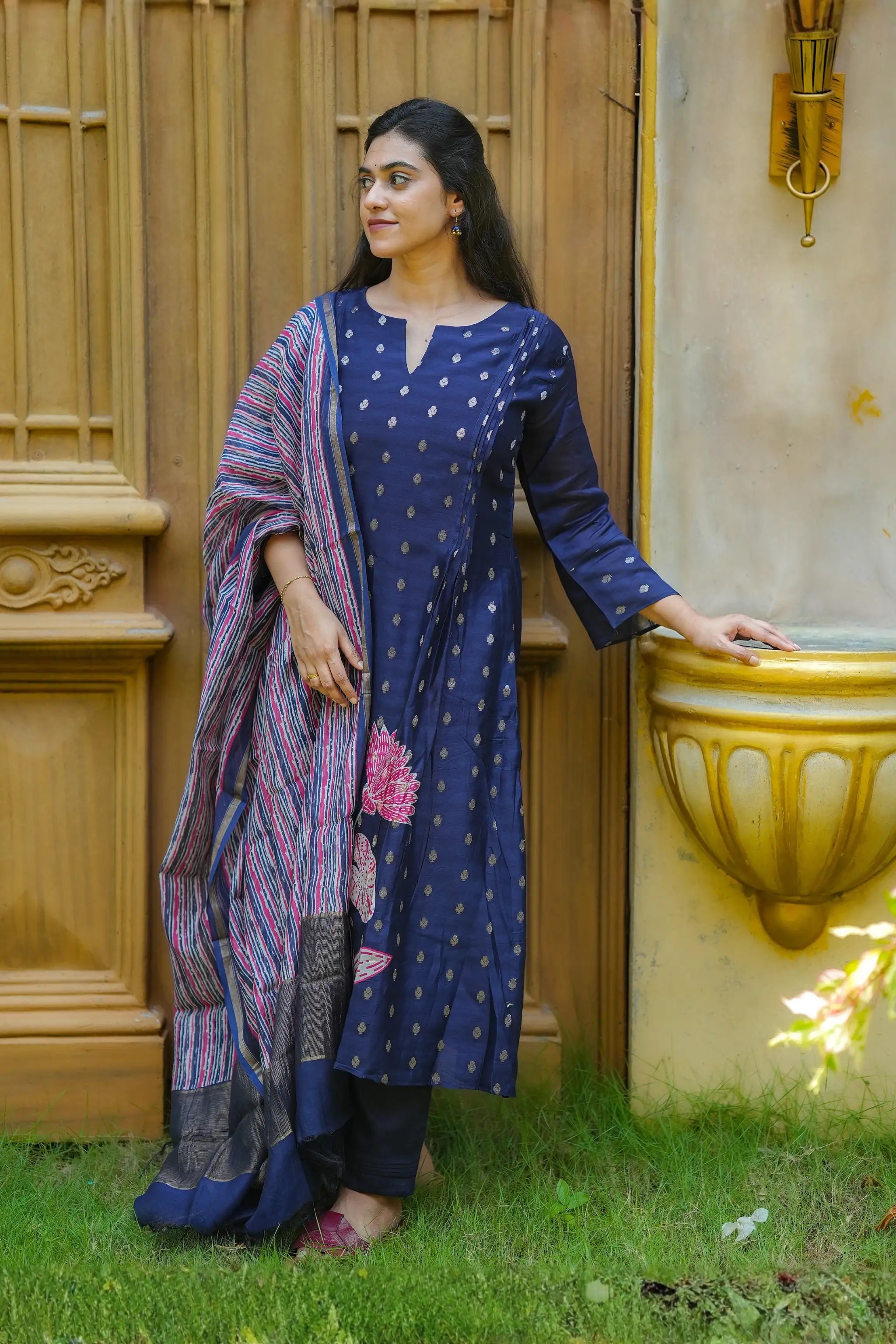 Woman in a blue traditional outfit standing outdoors next to a decorative wall.