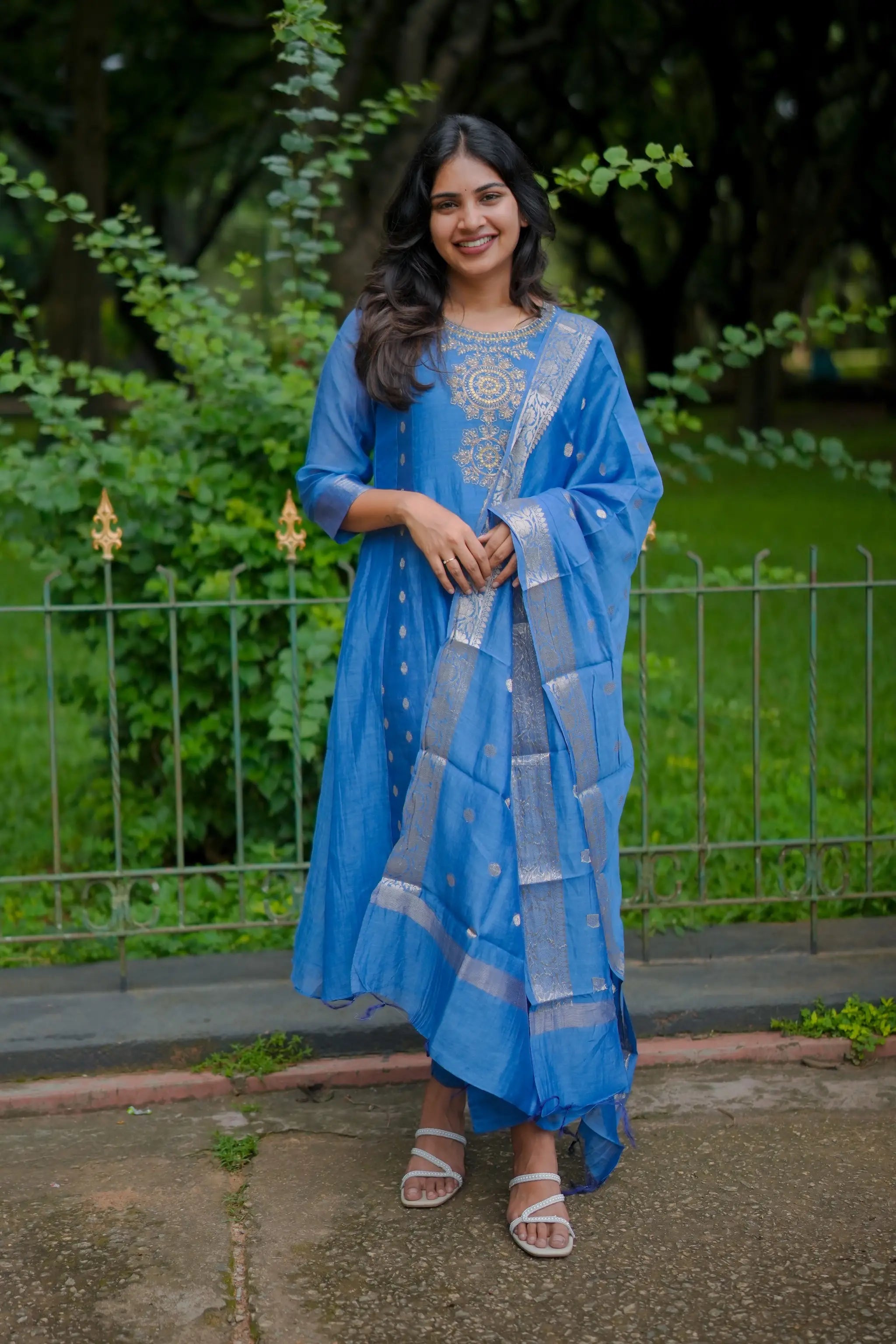 Woman in a blue traditional outfit standing outdoors with greenery in the background