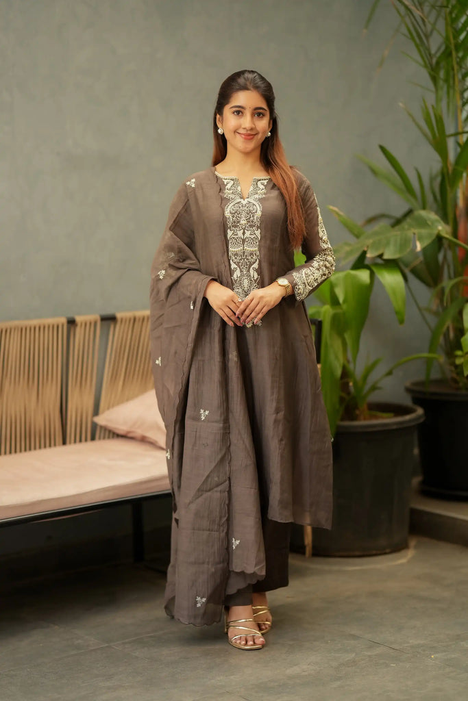 A woman models a traditional three-piece salwar kameez suit in taupe brown, featuring an embroidered kameez top with silver and white patterns, matching trousers, and a draped dupatta with small embroidered motifs, set against a modern indoor backdrop with a bench and potted plants.