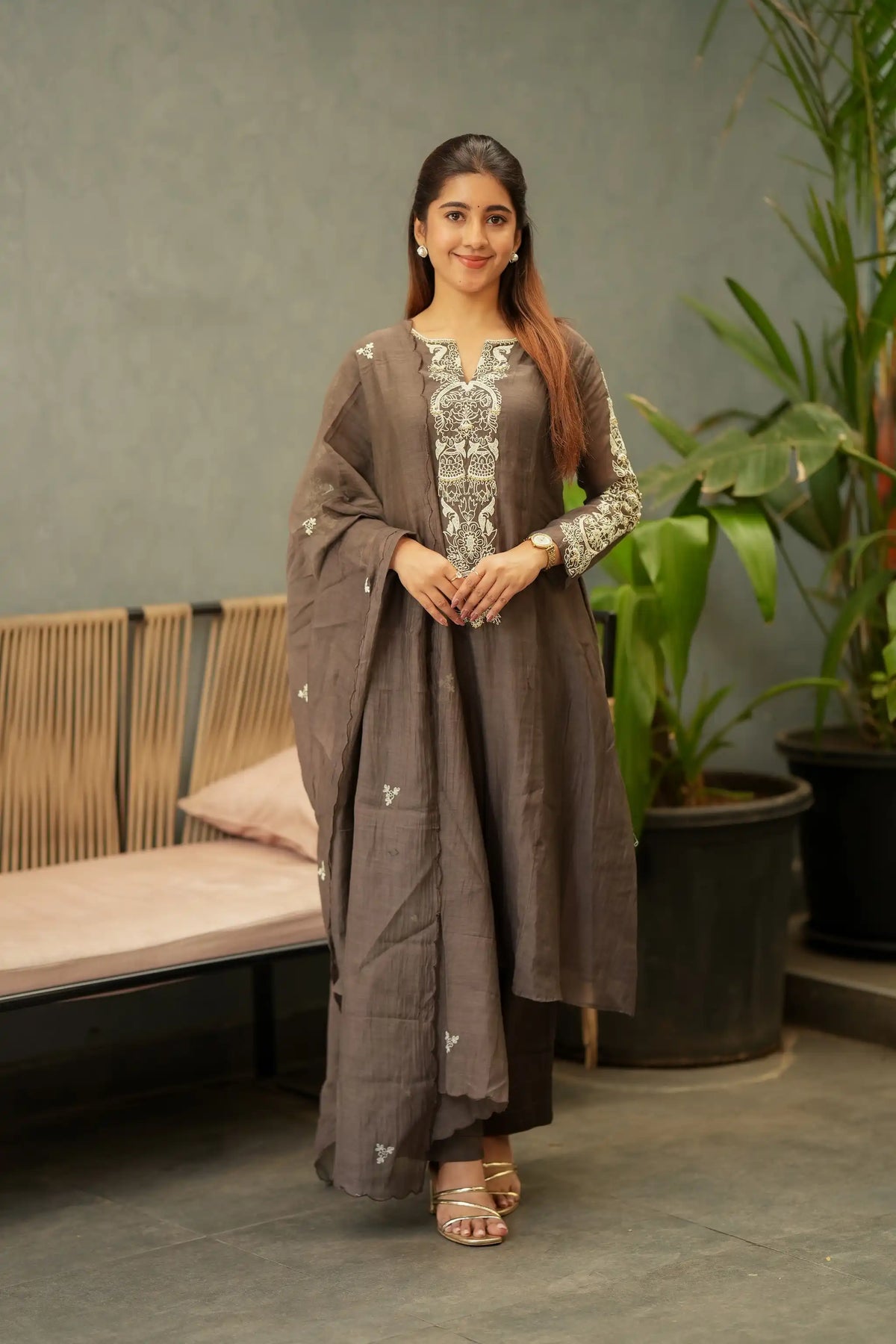 A woman models a traditional three-piece salwar kameez suit in taupe brown, featuring an embroidered kameez top with silver and white patterns, matching trousers, and a draped dupatta with small embroidered motifs, set against a modern indoor backdrop with a bench and potted plants.