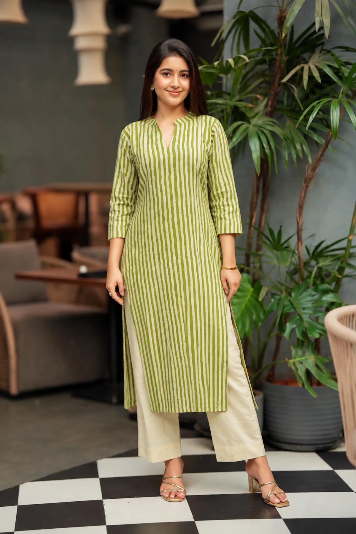 A woman modeling a light green and white vertically striped kurta with beige wide-leg pants, standing on a black and white checkered floor in a modern indoor setting with potted plants.