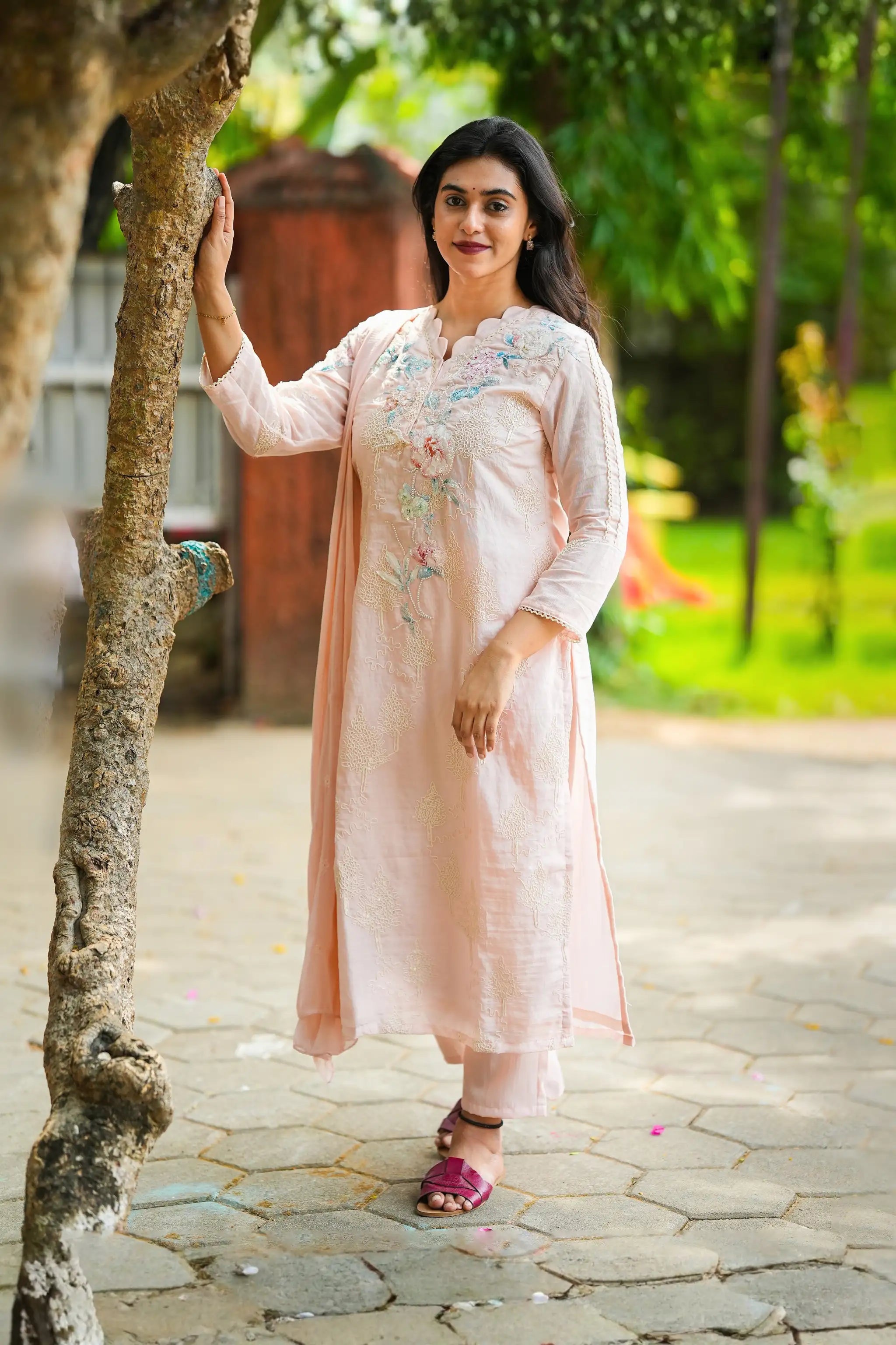 Woman in a light pink traditional outfit standing outdoors with greenery in the background