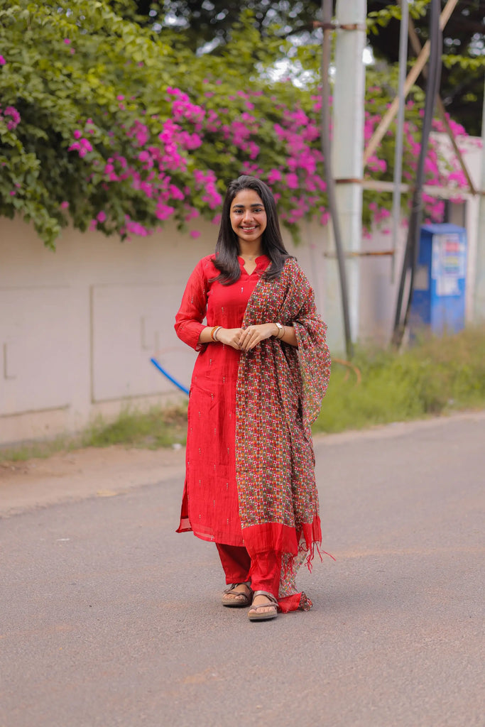 Woman in a red traditional outfit with a patterned shawl standing on a road with greenery and flowers in the background.