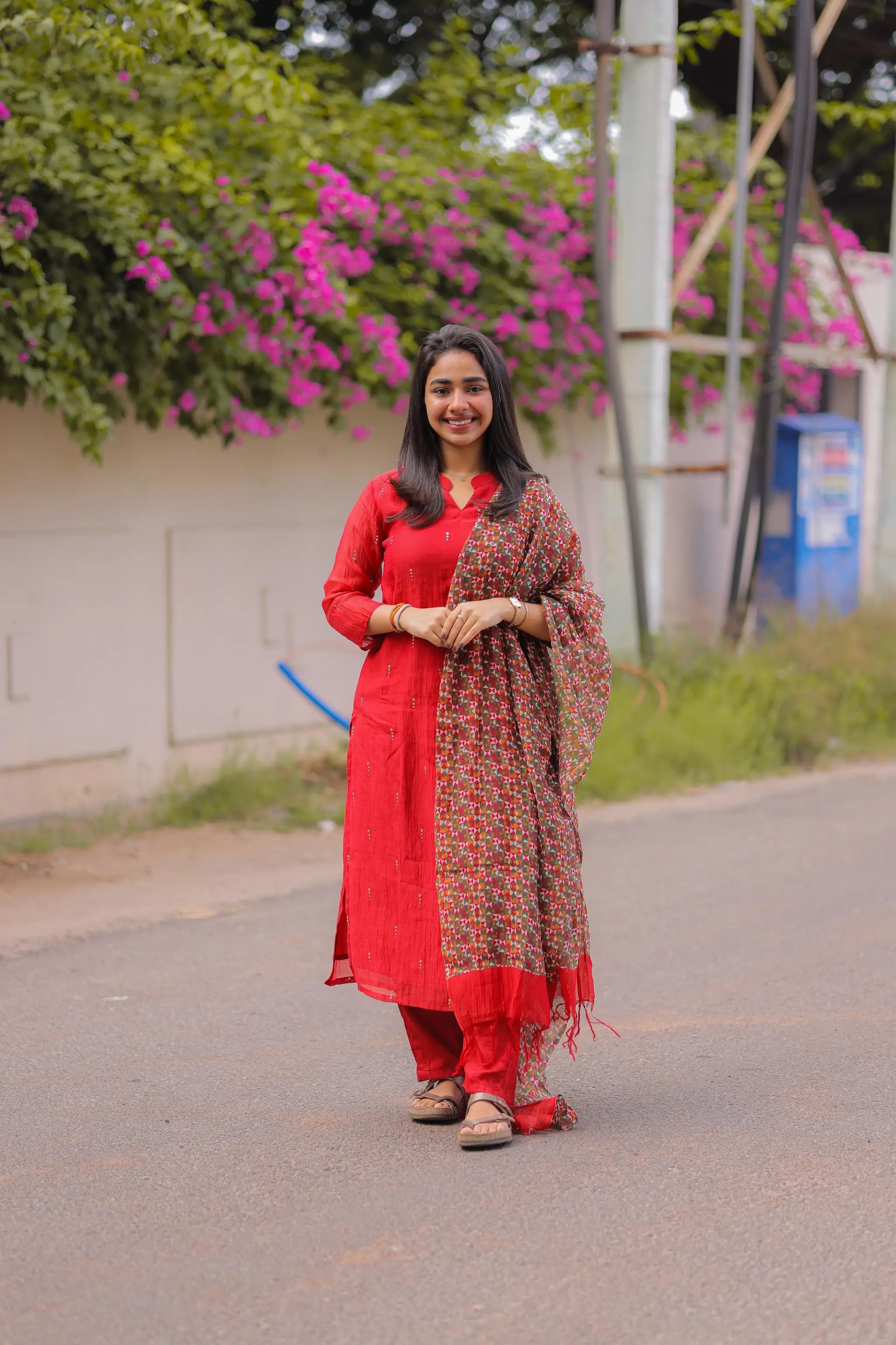 Woman in a red traditional outfit with a patterned shawl standing on a road with greenery and flowers in the background.
