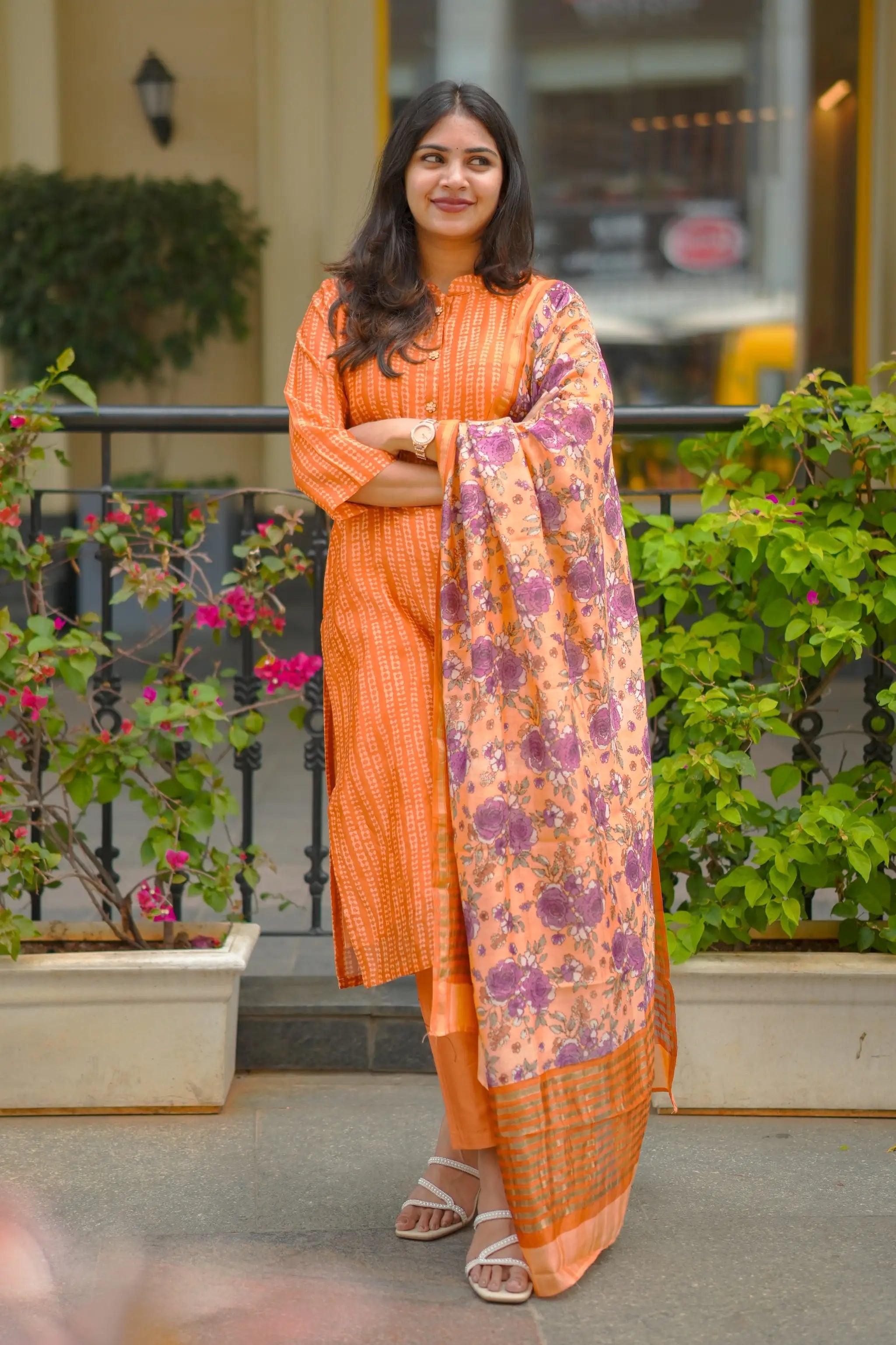 Woman in an orange traditional outfit with a floral dupatta standing on a balcony with plants and flowers.