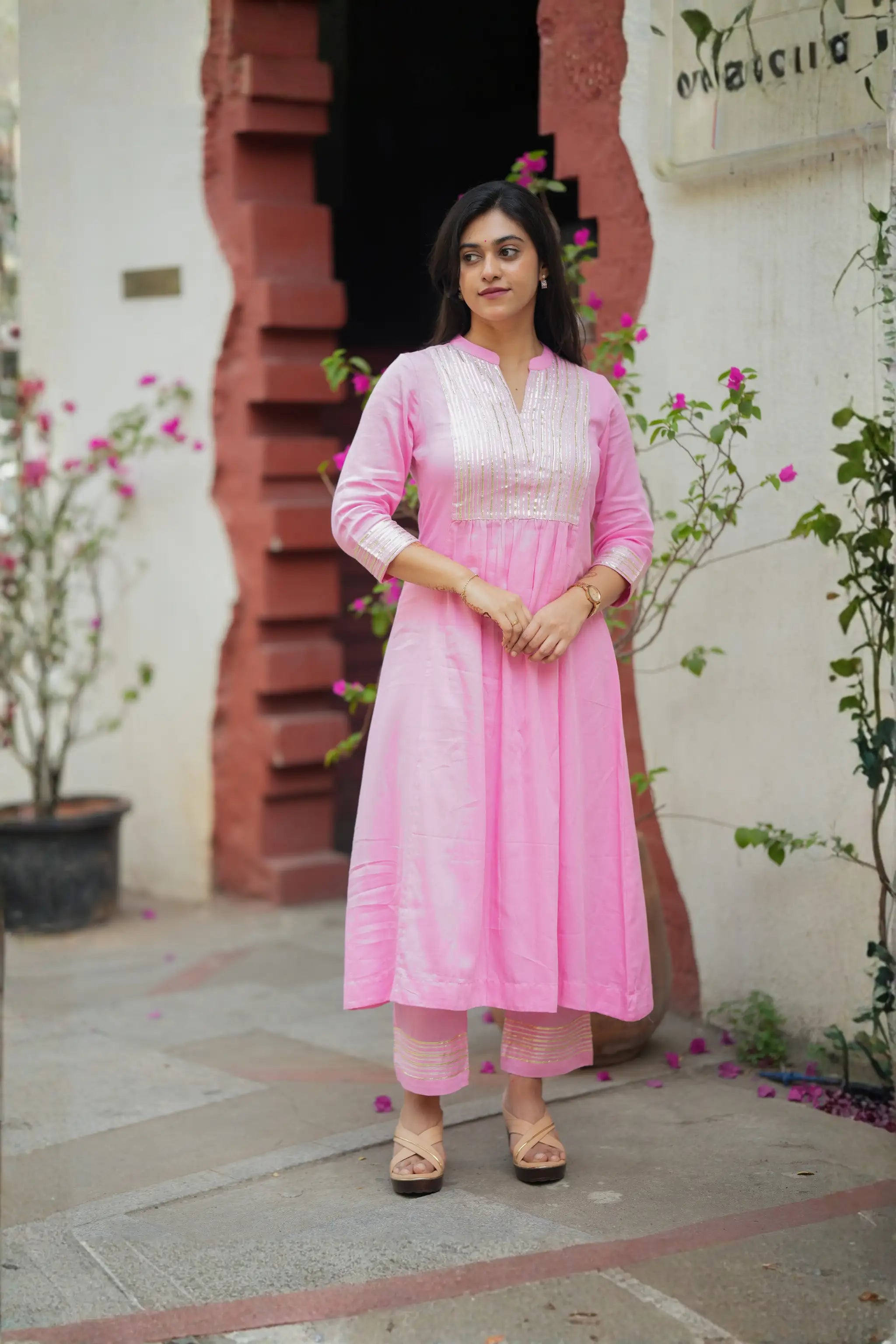 Woman in a pink traditional outfit standing in front of a red brick wall with plants.