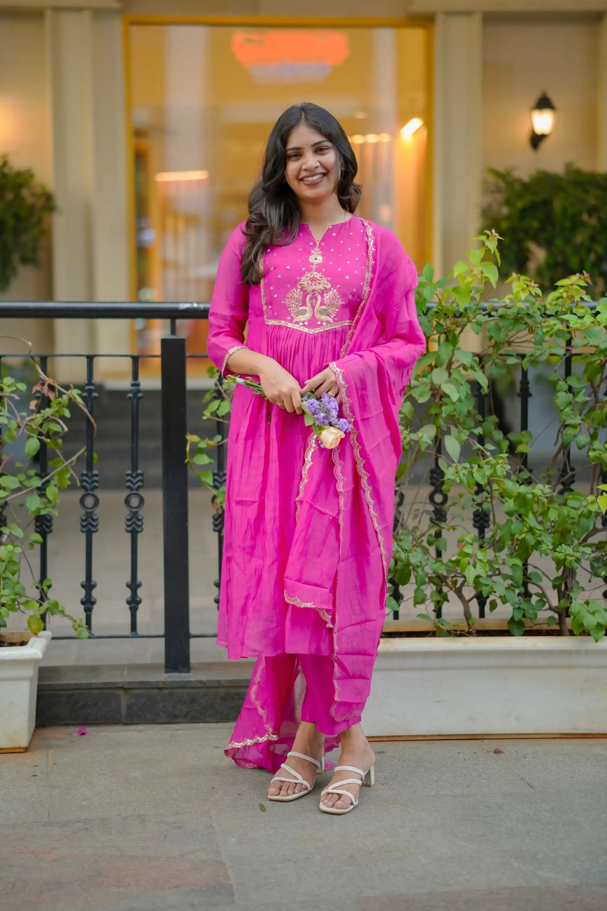Woman in a pink traditional outfit standing outdoors with greenery and a building in the background