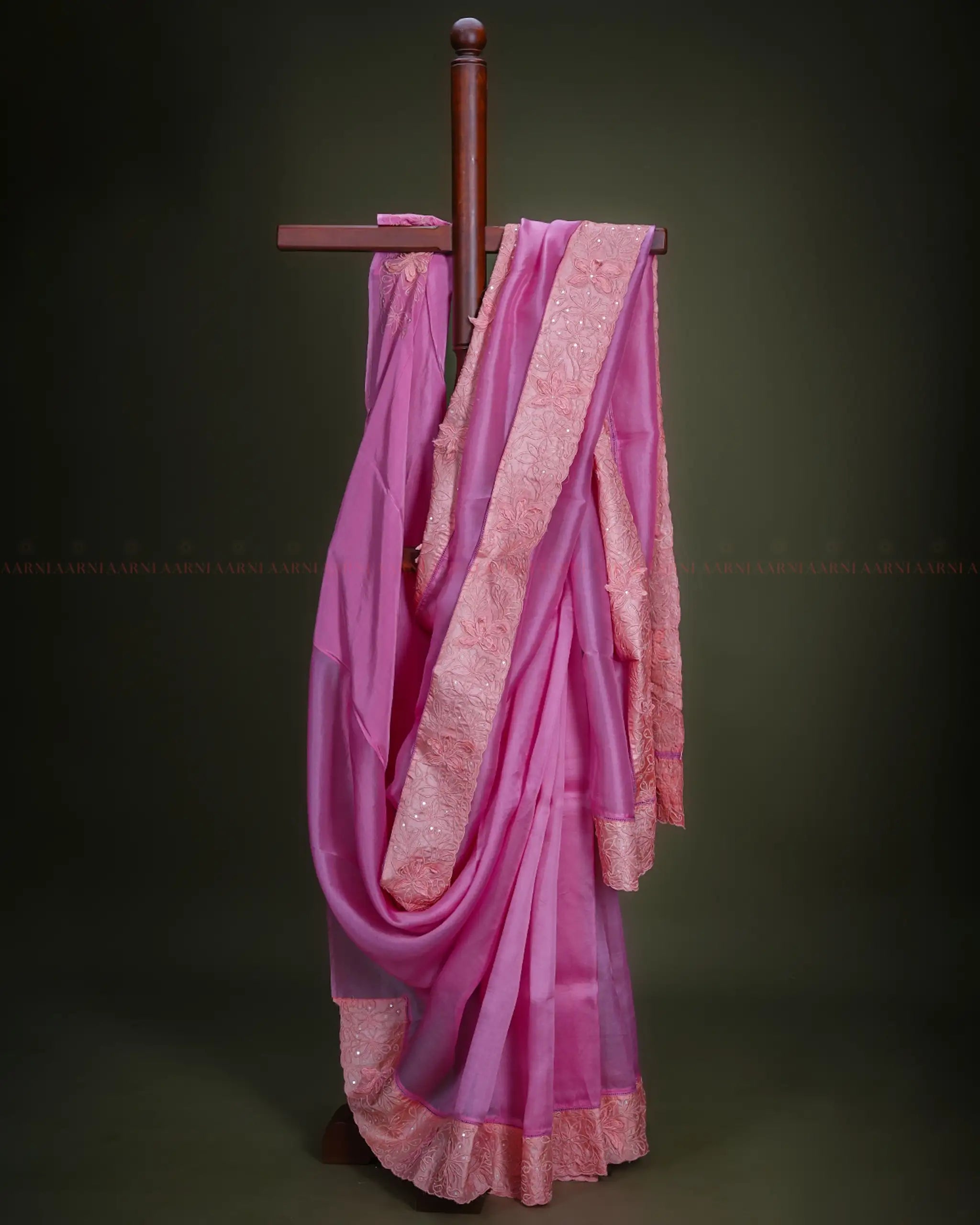 Pink organza saree with a border draped over a wooden rod against a dark background