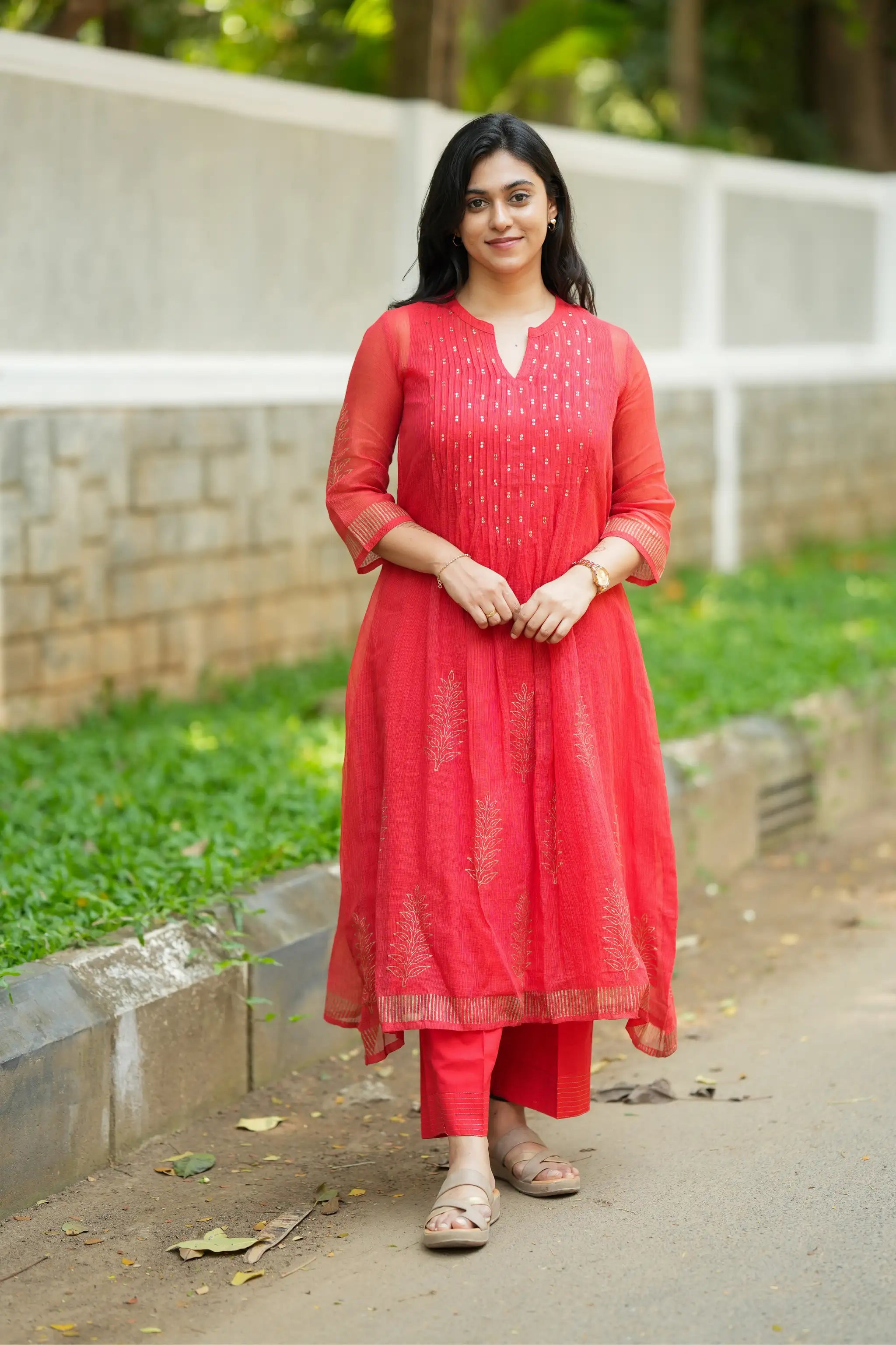 Woman in a red traditional outfit standing outdoors on a path with greenery.