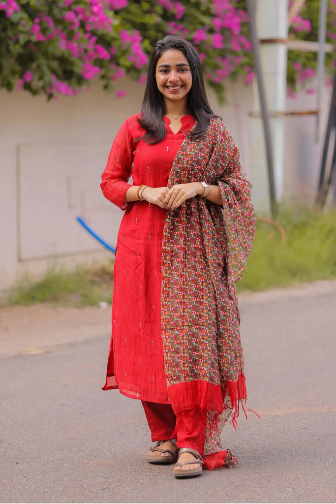 Woman in a red traditional outfit with a patterned scarf standing outdoors.