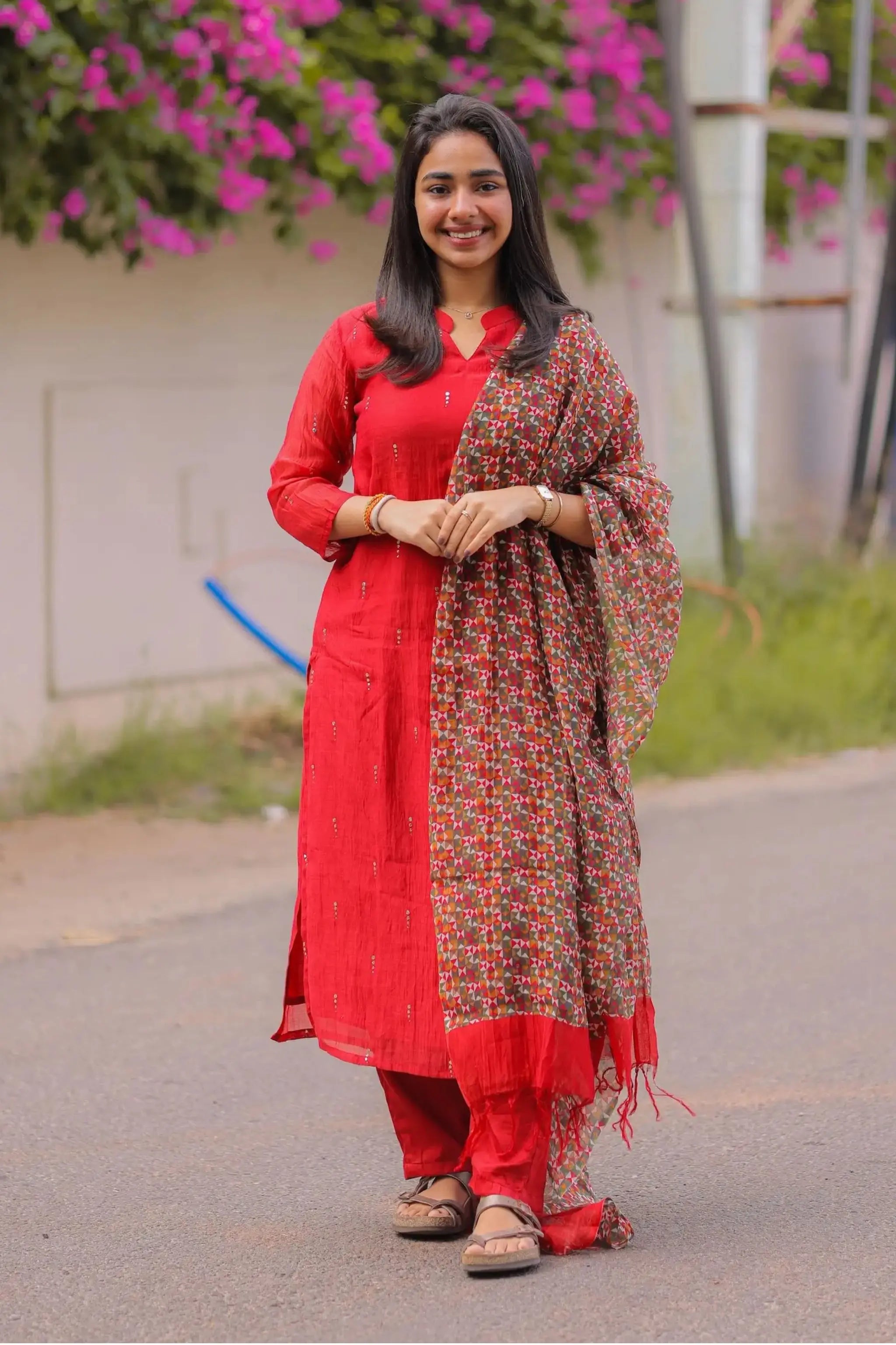 Woman in a red traditional outfit with a patterned scarf standing outdoors.