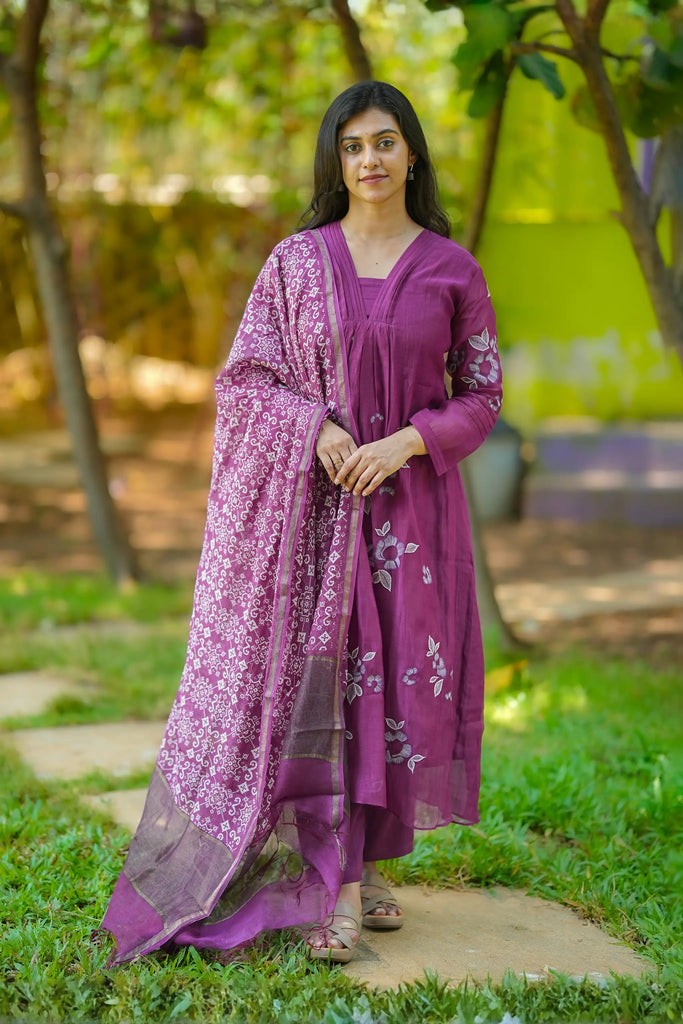 Woman in a purple traditional outfit standing outdoors with greenery in the background