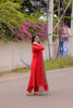 Woman in a red traditional outfit standing on a road with greenery and flowers in the background