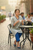Woman sitting at an outdoor cafe table holding a cup, with a blurred street scene in the background.