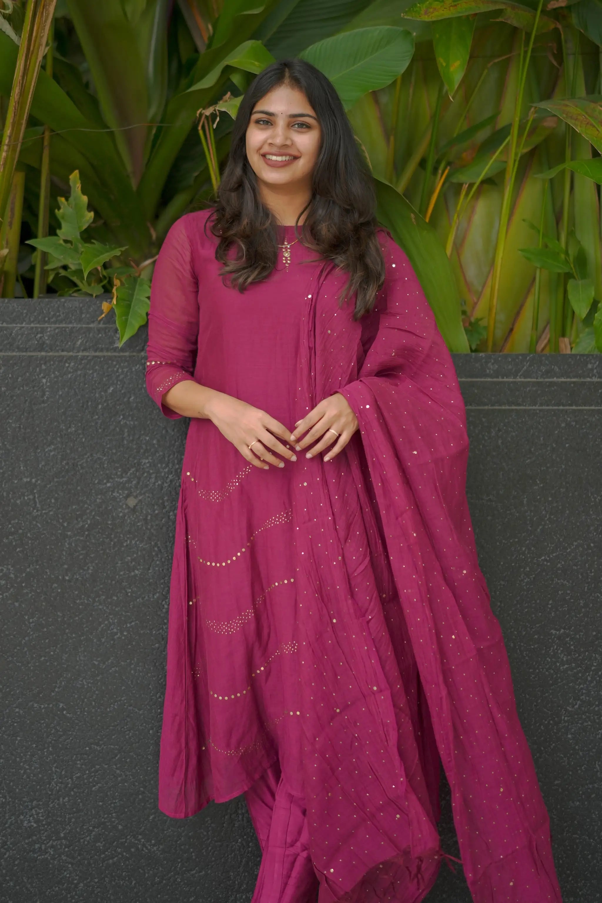 Woman in a pink traditional outfit standing against a dark wall with greenery in the background