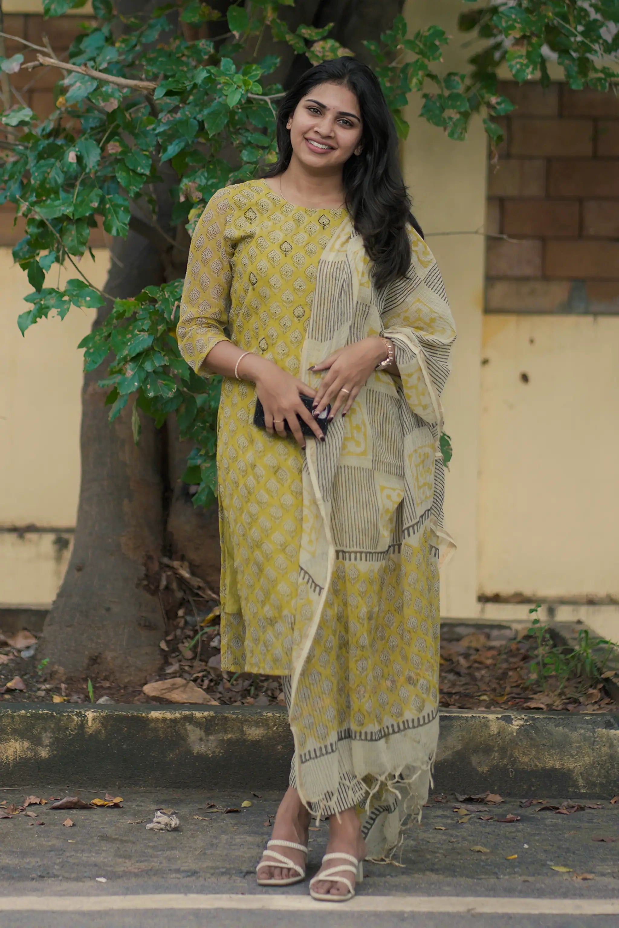 Woman in a yellow traditional outfit standing outdoors with greenery in the background