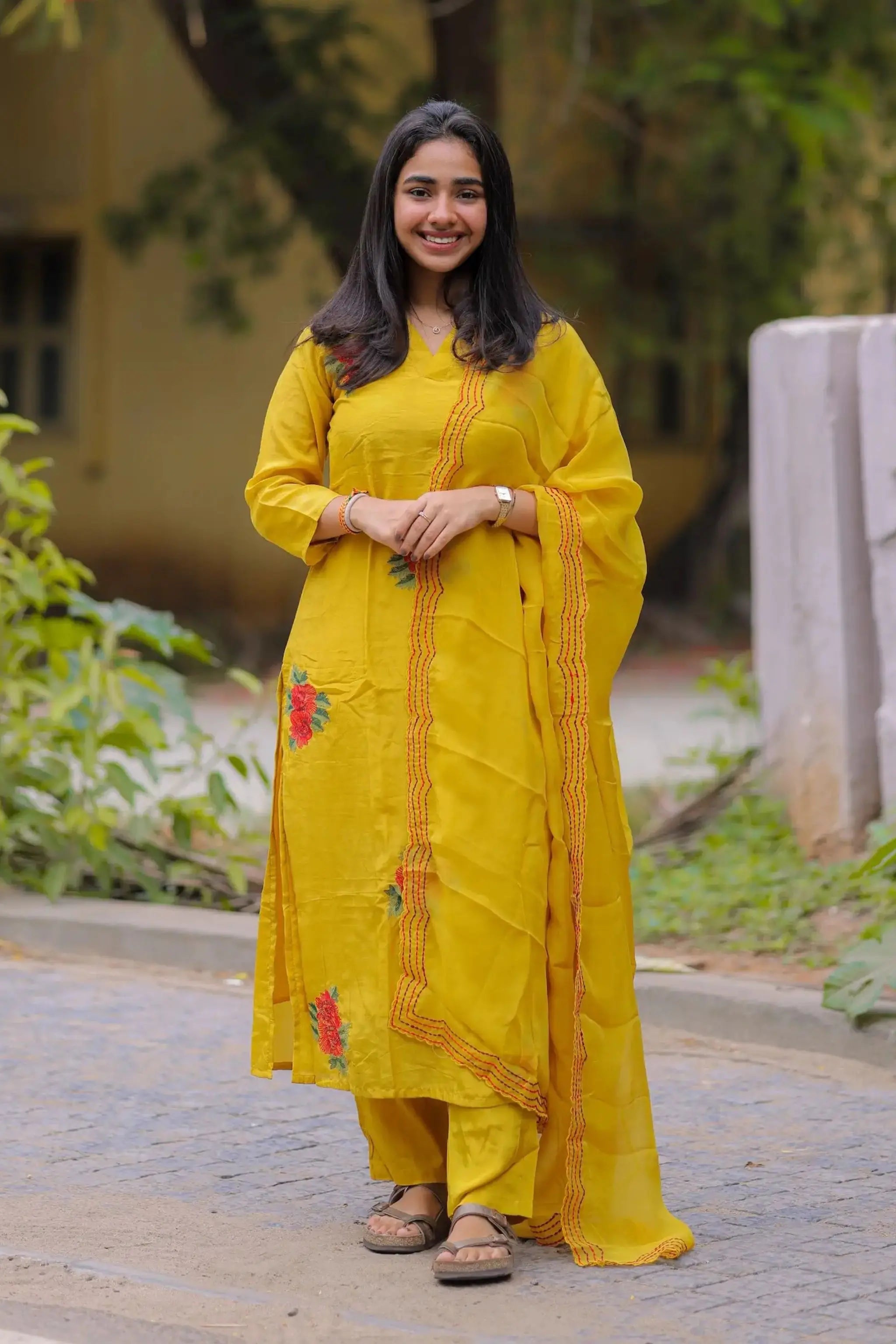 Woman in a yellow traditional outfit standing outdoors with greenery in the background