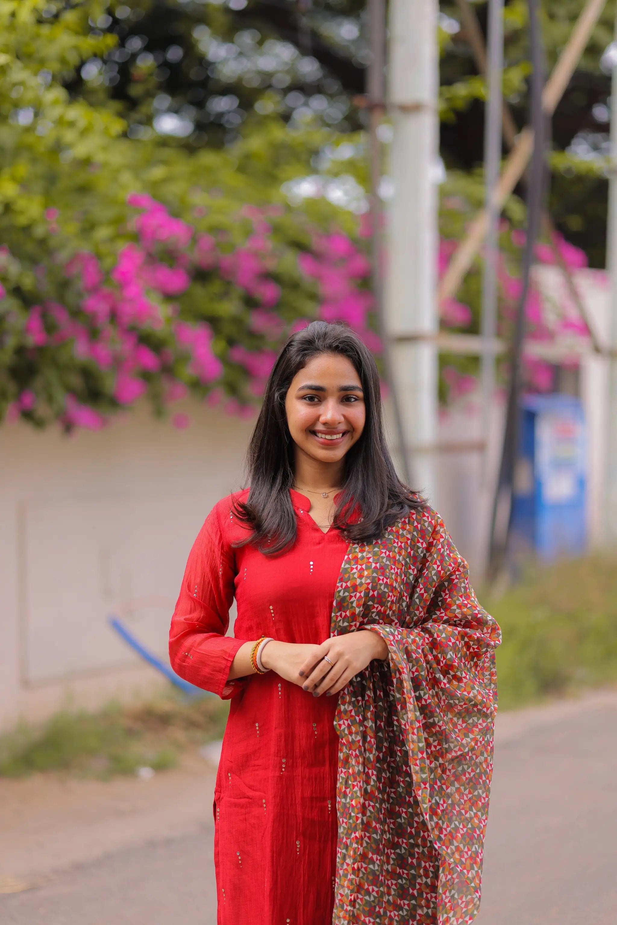 Woman in a red outfit with a patterned scarf standing outdoors with greenery and flowers in the background